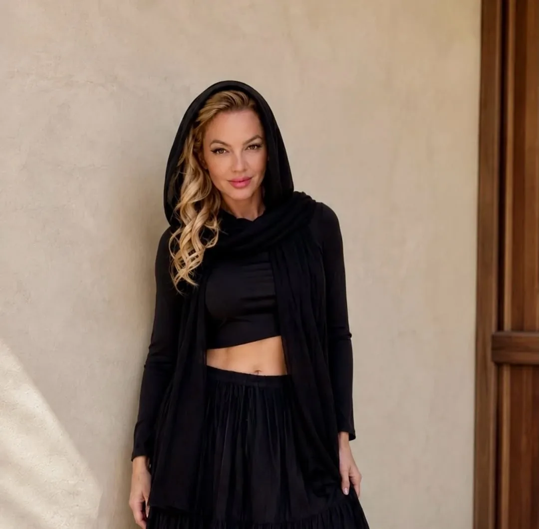 Roberta Graciana de capuz preto e top, encostada em uma parede clara