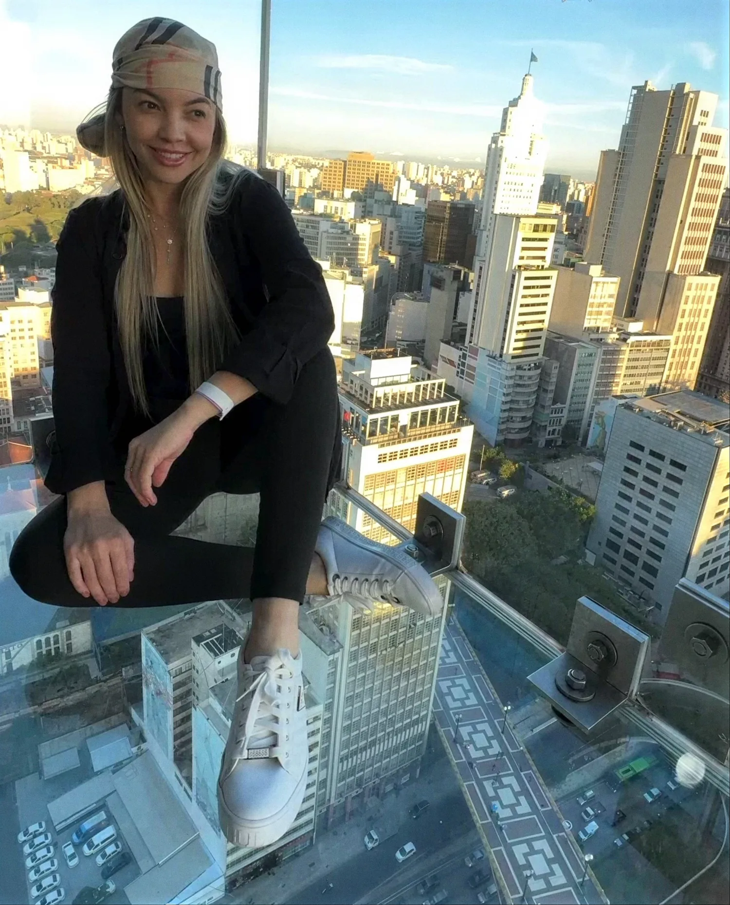 Roberta Graciana agachada em um mirante de vidro, com a cidade de São Paulo ao fundo